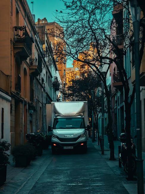 A white Iveco moving van parked on a narrow urban street between tall buildings with residential balconies and window shutters. The van's cargo area is covered with a white protective wrapping, indica