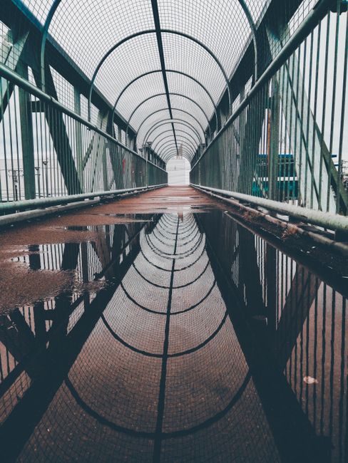 Inside a covered outdoor pedestrian bridge with a curved, transparent roof and metal railing on both sides. The walkway surface is wet, showing puddles that reflect the bridge's structure. The bridge leads toward an opening with visible sky beyond, suggesting an outdoor connection. This scene depicts a pathway that could be used during a property move for access between parts of a residence or building. Occasionally, Barnes Removals assists with home relocations involving careful transport through such access points, ensuring furniture and boxes are moved safely from inside the property onto a van. The bridge's design and environment indicate a location that might be part of a larger estate or urban setting commonly used during home removals or furniture transport processes, supporting efficient logistical planning for removals and packing through external access routes.