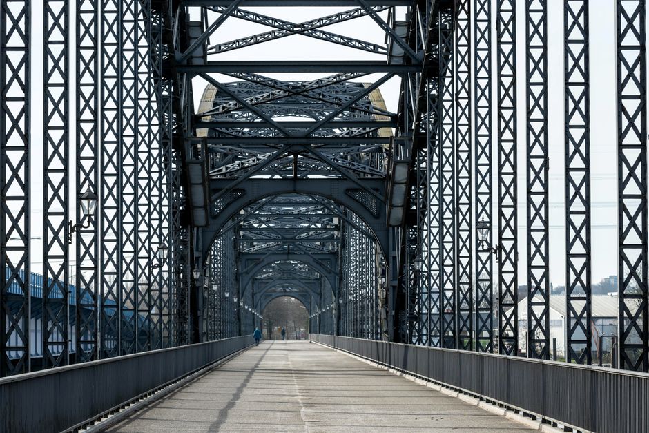 A long view of a metal lattice bridge structure with an arched design, featuring steel framework with crisscrossing beams and vertical supports, extending over a pathway. The pathway inside the bridge is paved, with a couple of pedestrians walking in the distance. On the right side, a black van is parked, partially visible behind the railing. The bridge appears to be used for foot and vehicle traffic, situated in an urban or semi-industrial environment. Natural daylight illuminates the scene, highlighting the geometric design of the steel framework. This setting captures a typical scene during a home relocation or moving process involving transportation over the bridge, demonstrating the logistics of furniture and belongings being transferred through such structures, aligned with the services provided by Barnes Removals.