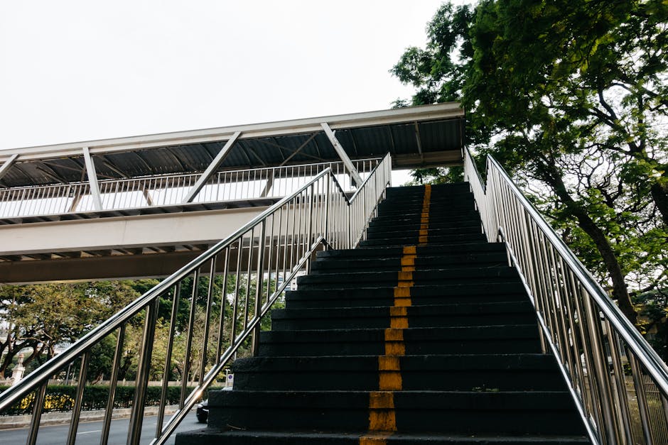 Inside a covered outdoor pedestrian bridge with a curved, transparent roof and metal railing on both sides. The walkway surface is wet, showing puddles that reflect the bridge's structure. The bridge leads toward an opening with visible sky beyond, suggesting an outdoor connection. This scene depicts a pathway that could be used during a property move for access between parts of a residence or building. Occasionally, Barnes Removals assists with home relocations involving careful transport through such access points, ensuring furniture and boxes are moved safely from inside the property onto a van. The bridge's design and environment indicate a location that might be part of a larger estate or urban setting commonly used during home removals or furniture transport processes, supporting efficient logistical planning for removals and packing through external access routes.